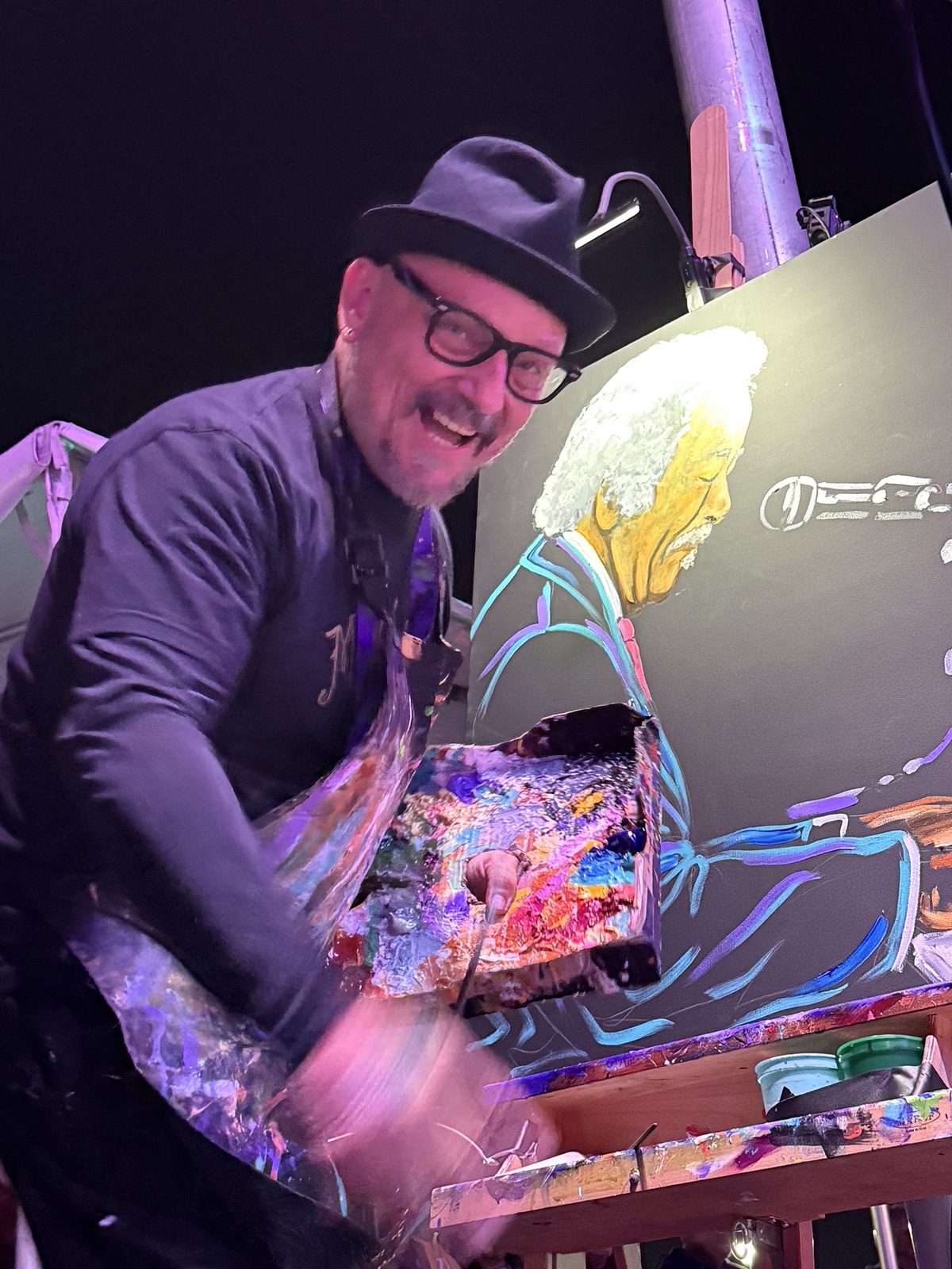 Frenchy grinning at his easel on the NOLA Funk Fest main stage, palette in hand, with his Allen Toussaint painting in progress behind him
